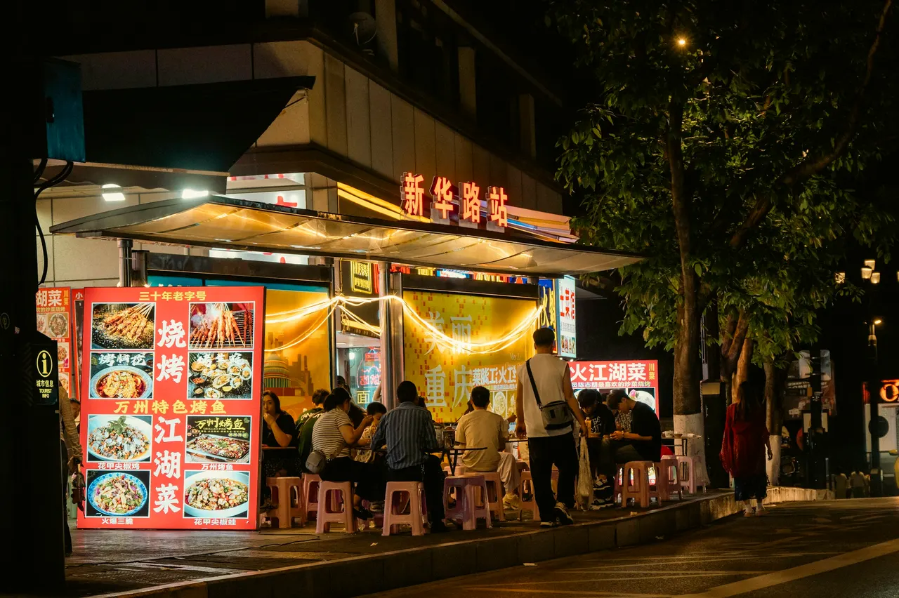 Outdoor dining table on the pavement beside an illuminated photo menu board at night.