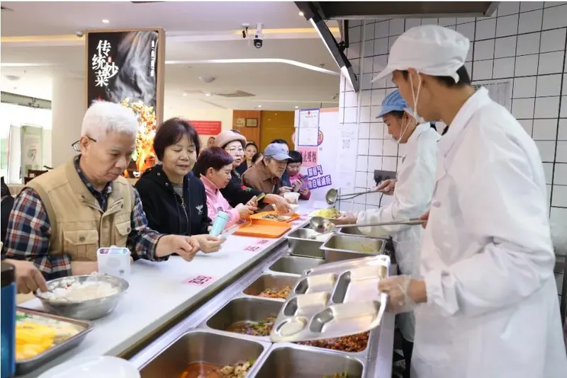 Residents choosing dishes at a community canteen counter, with staff serving from metal food trays.