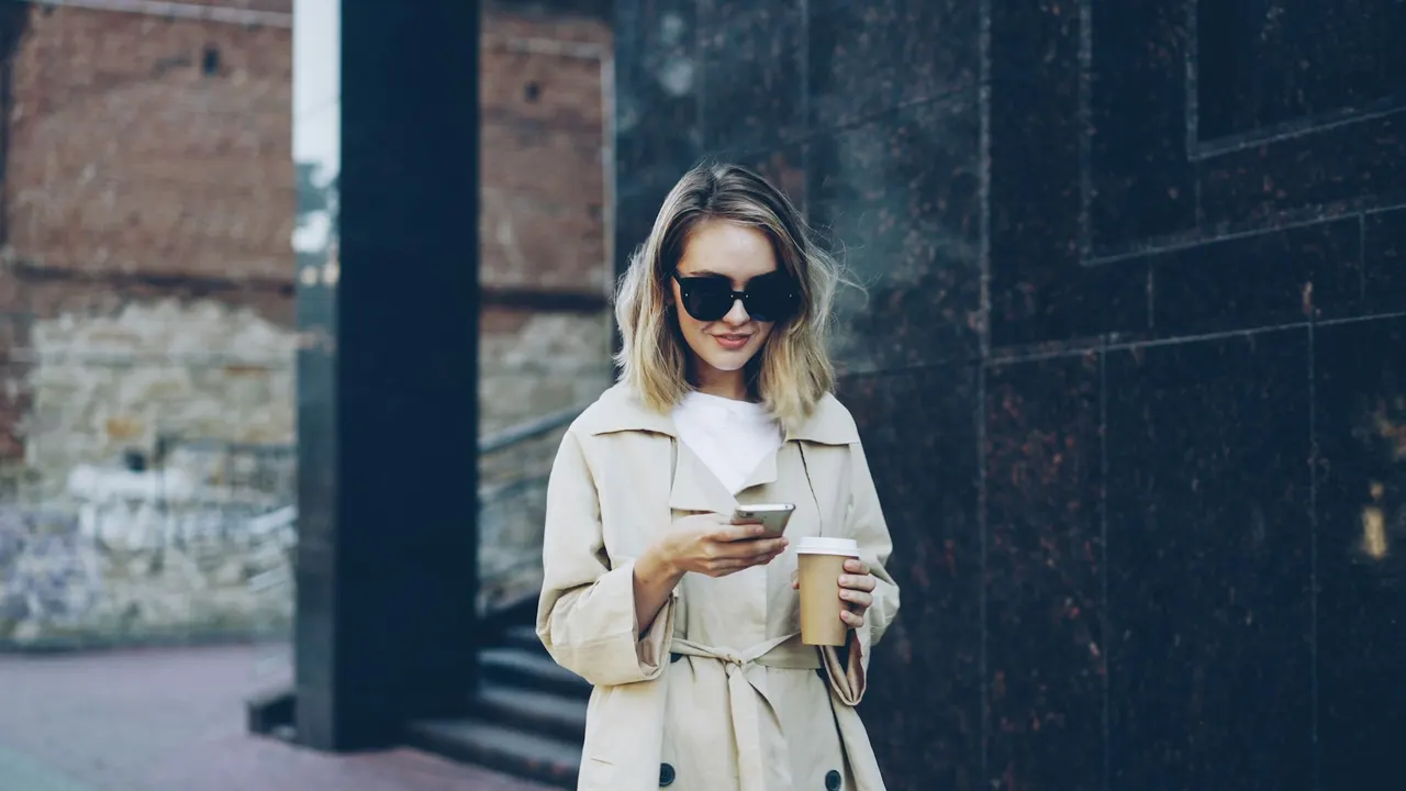 Một phụ nữ nhìn điện thoại và cầm cà phê mang đi trên đường phố
