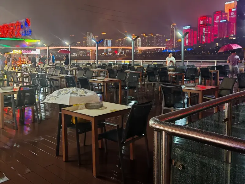 Tables and chairs on an open-air boat deck, with a railing and Chongqing’s illuminated riverfront behind them
