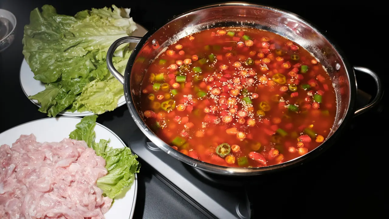 A pot of red broth beside plates of lettuce and sliced meat on a dark table