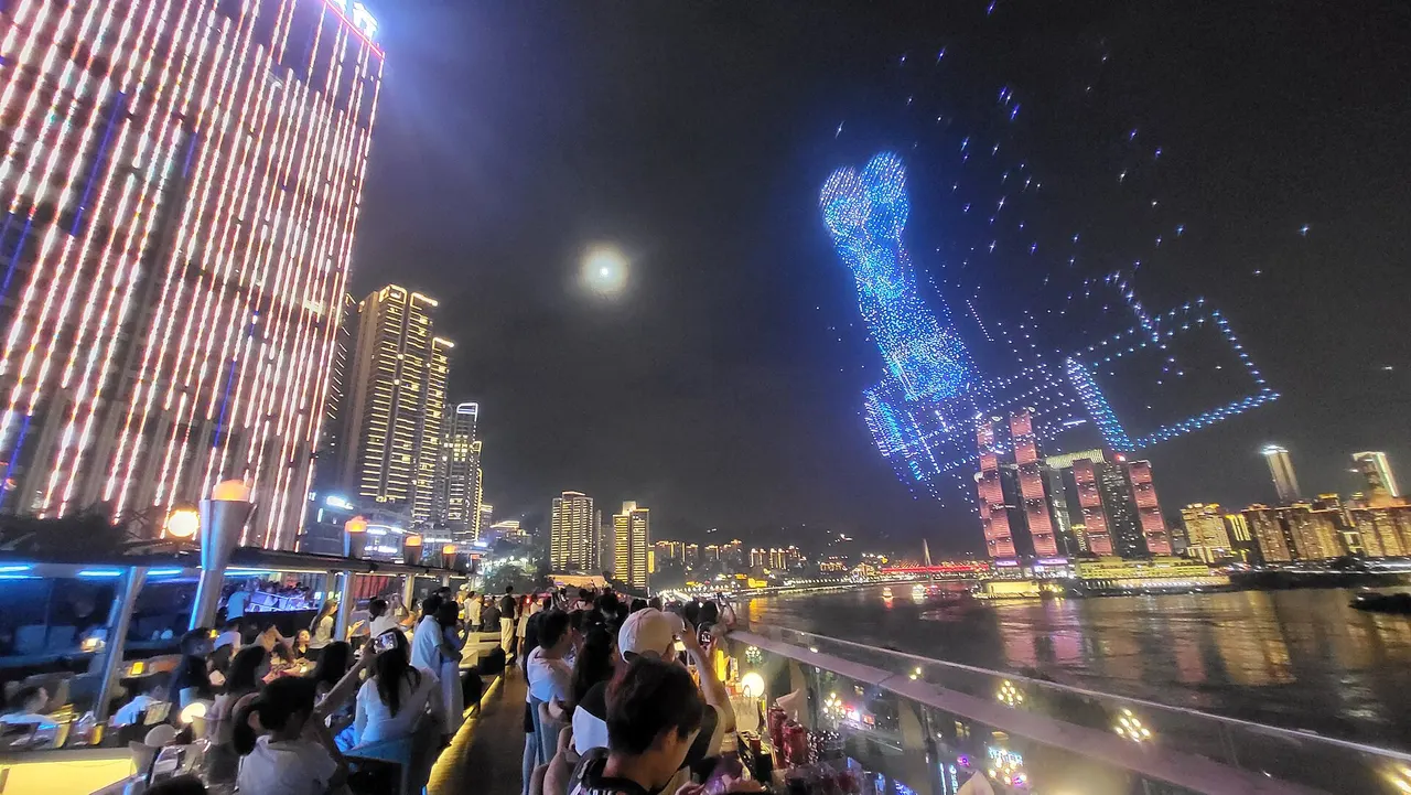 Patrons on a riverside terrace watching a drone light display across the river.