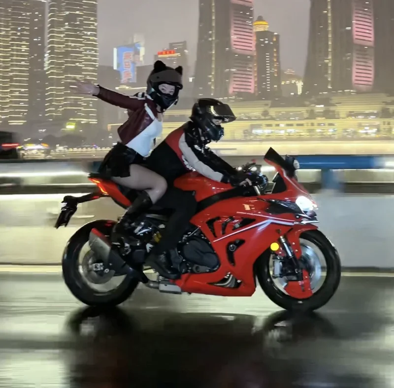 Duas pessoas de capacete em uma moto vermelha, com prédios iluminados de Chongqing ao fundo à noite