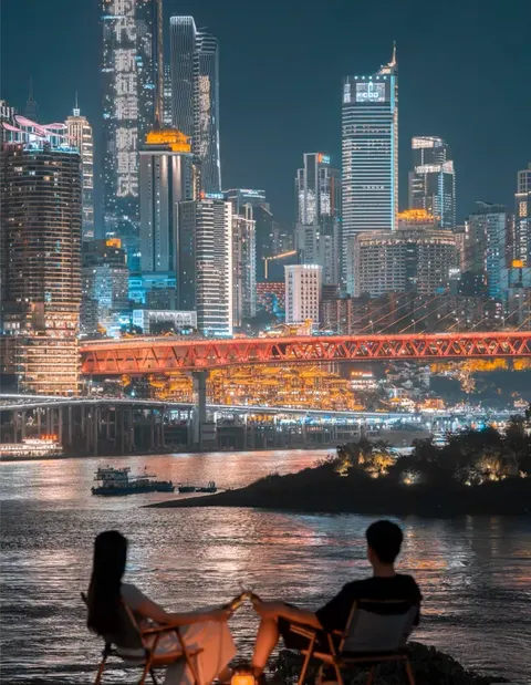 Couple in everyday clothes holding hands on riverside folding chairs beside a lantern, with Hongya Cave and Qiansimen Bridge lit behind them