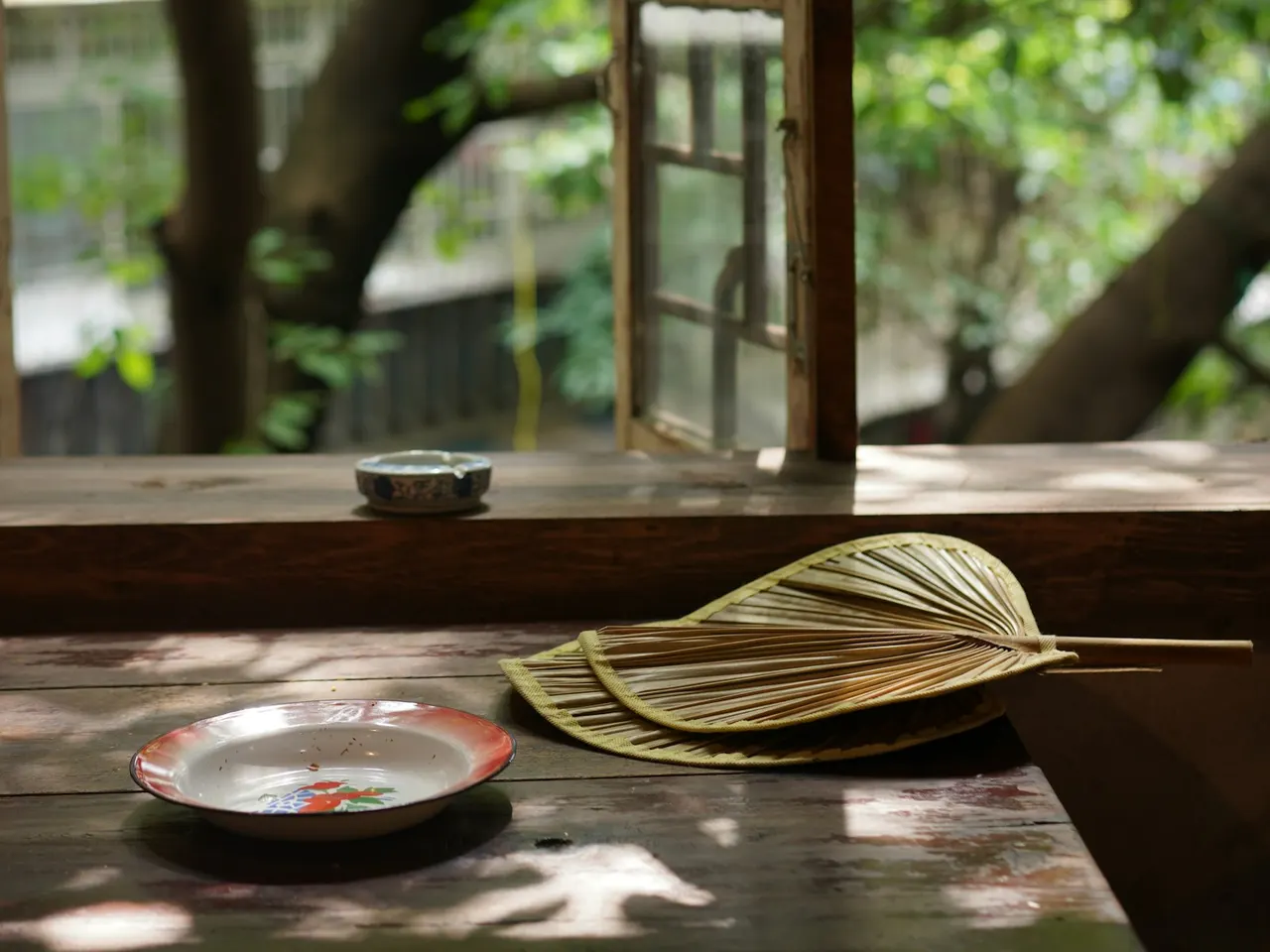 Woven hand fan and saucer on a wooden table beside an open teahouse window in Mountain City Lane