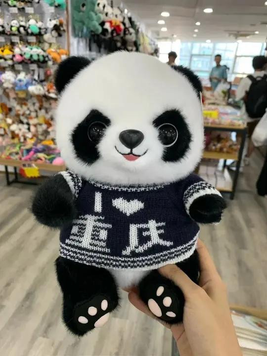 A hand holds a panda plush wearing a dark sweater with a heart and Chongqing lettering.