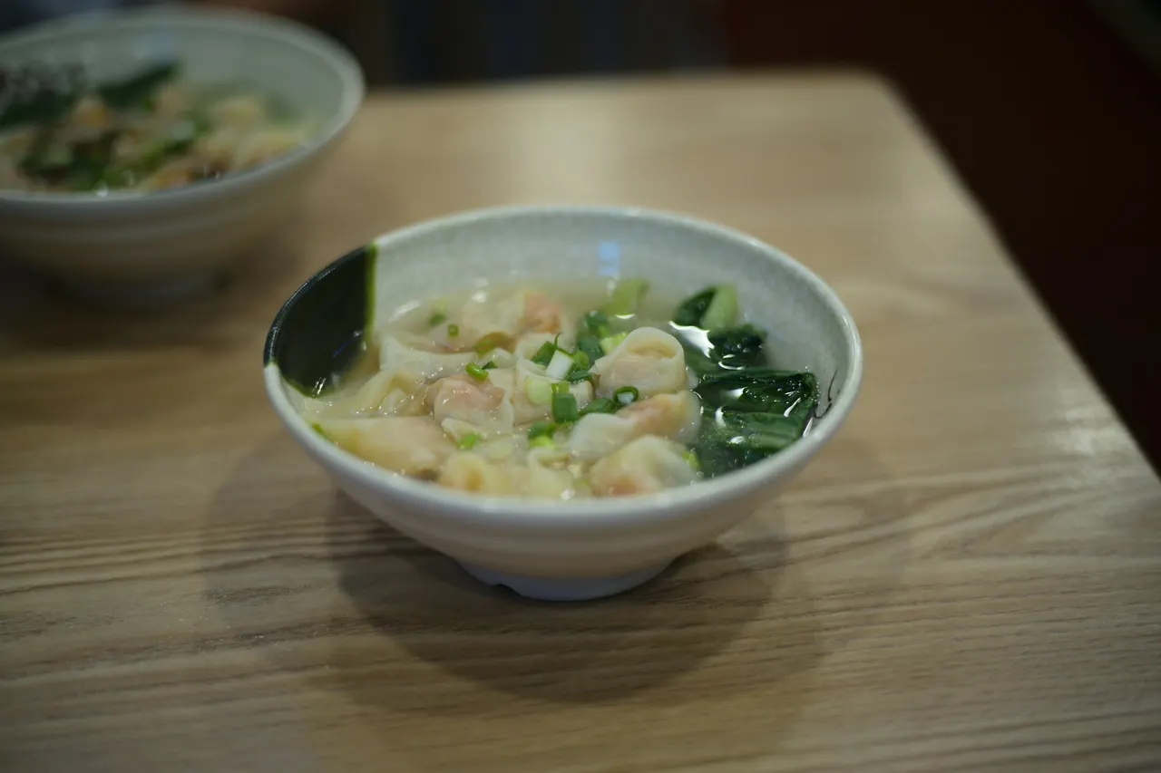 A bowl of wonton soup with leafy greens and spring onions on a restaurant table