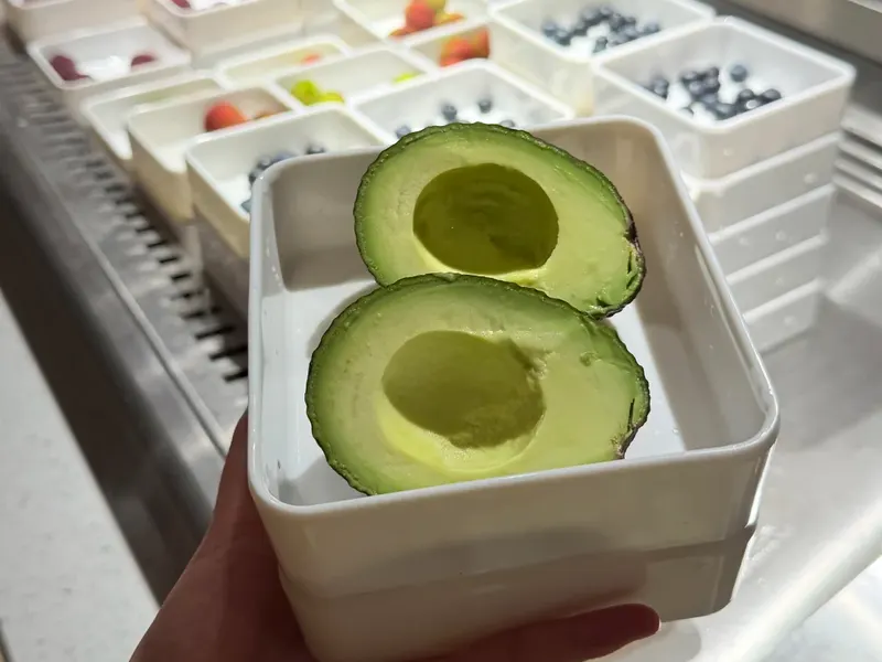 A hand holds a white bowl with two avocado halves in front of the refrigerated fruit counter at Qianxi