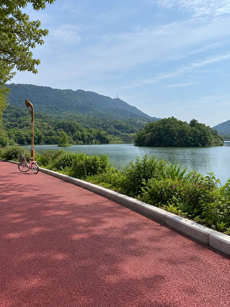 Red path beside Xuantian Lake, with a pink bicycle farther along the shore and wooded hills across the water