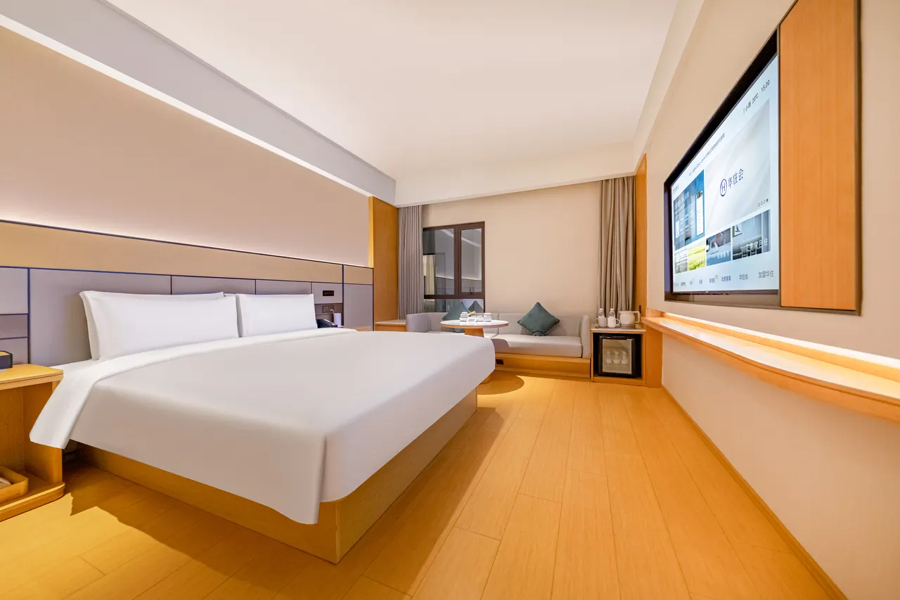 White king bed and pale wood furniture, with a small sofa near the window and a wall-mounted television.